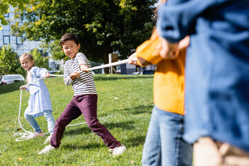 Fototapeta premium Asian boy playing tug of war with friends on blurred foreground on grass in park