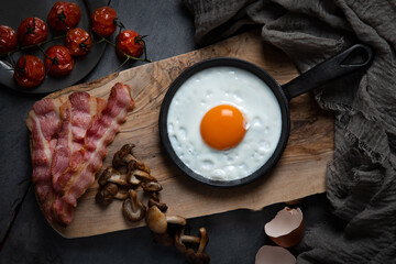 Overhead of fried egg in a pan on a wooden board, with cooked bacon, mushrooms and tomatoes arranged around, against a dark background.