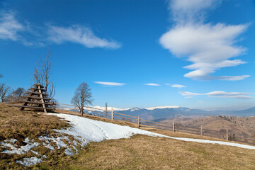 Spring in the mountains, wide-angle landscape, pasture with withered grass and melting snow, village on the distant slope, snowcapped mountains on the horizon, bright blue sky. Carpathians.