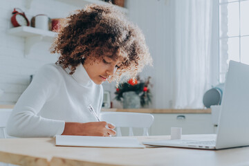 Student girl with trendy kinky hair sitting against notebook and writing notes in copybook enjoying...