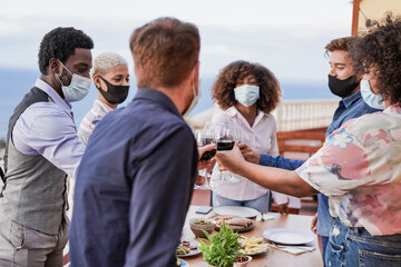 Naklejka premium Young multiracial friends enjoy dinner at home on patio and cheering with wine while wearing protective face mask for coronavirus