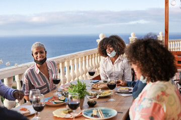Happy multiracial friends enjoy dinner together outdoor while wearing surgical face mask under chin