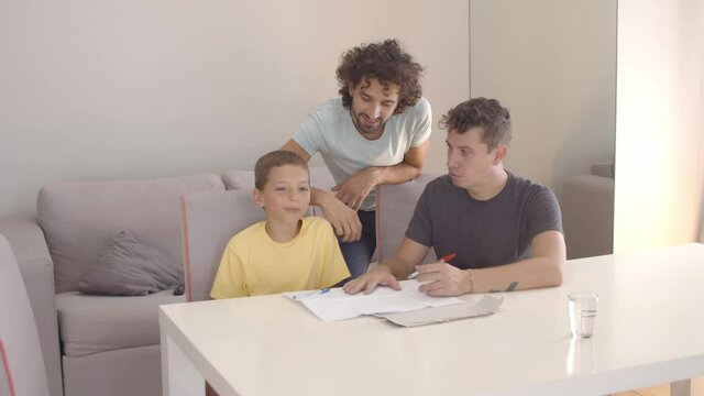 Gay parents helping son with writing task, sitting at table with papers, speaking and holding pens. Static shot. Family and parenthood concept