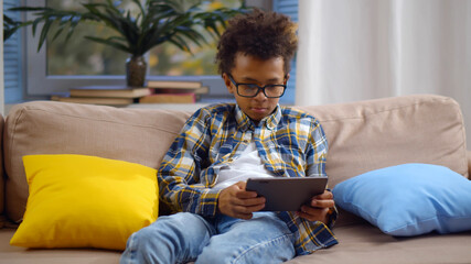 Cute african child in glasses sitting on sofa playing game on digital tablet.