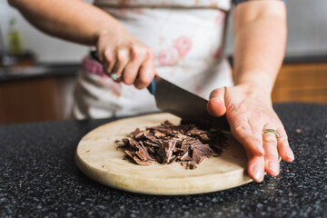 Woman chopping chocolate on wooden kitchen board in the kitchen wearing apron. Preparing ingredients for baking.