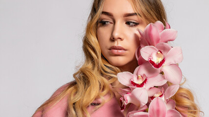 Seductive blonde woman in pink jacket posing in studio on grey background. elegant model in pastel casual spring outfit. Beautiful girl with healthy skin and branch orchid. 16:9 panoramic format.