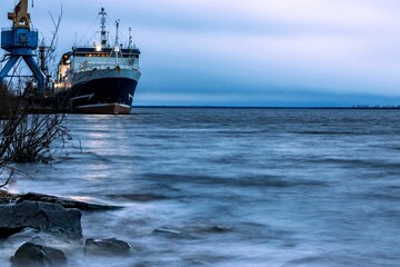 A seagoing vessel with glowing searchlights is moored at the pier on a large river. Morning twilight. Sea freight concept