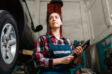 Portrait of a young caucasian female mechanic, in uniform, with a tablet in her hands. The wheel of the car on the right. Bottom view