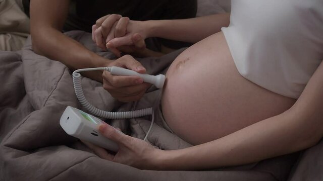 asian pregnant woman holding pocket fetal doppler to monitor baby heart beat. expectant mother touching belly. parent care future baby. father scan mother belly.