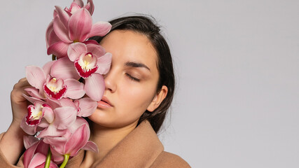 Seductive woman in brown jacket posing in studio on grey background.elegant model in pastel casual spring outfit. Beautiful girl with healthy skin and branch orchid. 16:9 panoramic format.