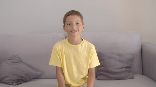 Cheerful positive boy wearing yellow t-shirt, sitting on couch at home, keeping hands on lap, looking at camera and smiling. Front view. Kids portrait or childhood concept