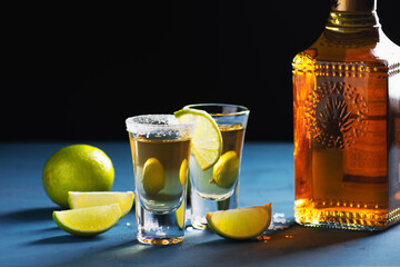 A bottle of golden mexican tequila and shots with lime and salt stand on a blue background,...