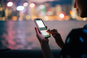 Fototapeta na wymiar Crop young woman messaging on smartphone with blank screen on blurred night city background