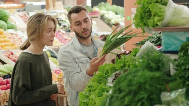 Handheld medium shot of cheerful young couple shopping for fresh produce at vegetable stand at market. They are putting scallions into paper bag and chatting