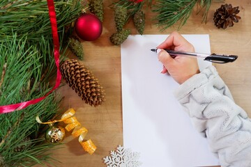 A girl writes a New Year's greeting card or Christmas wish sheet on a white piece of paper. Background of Christmas tree toys and evergreen pine needles