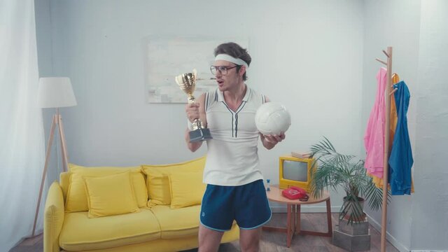 Sportsman holding trophy cup and sports ball, while standing at home