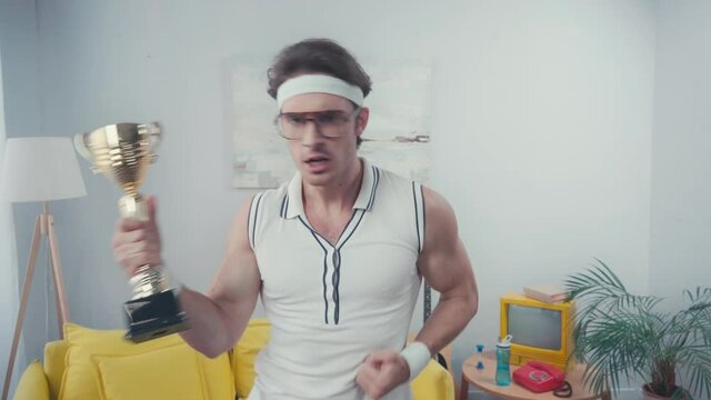 Sportsman with yeah gesture, celebrating win, while holding cup trophy at home