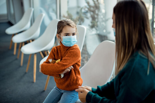 Small Girl With Face Mask Pouting While Being With Her Mother At Hospital's Waiting Room.