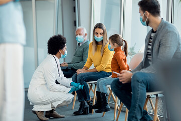 Mother and daughter talking to African American doctor while wearing face masks at hospital waiting room.