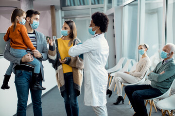 African American pediatrician talking to young family in a hallway at medical clinic during coronavirus pandemic.