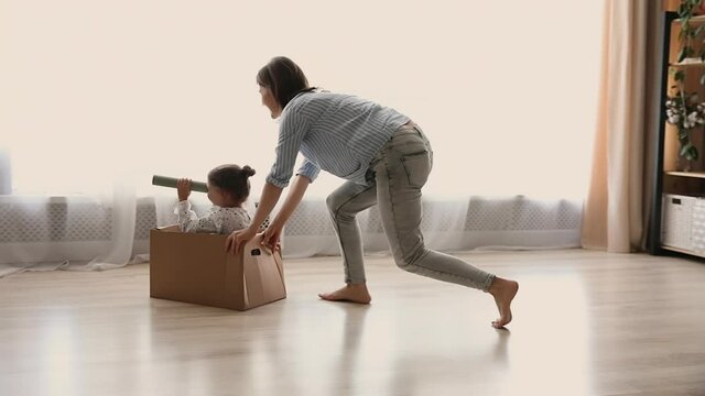 Mother play with little daughter rides her while she sitting in carton box, kid look in distance through paper tube imagines herself discoverer have fun at warm living room. Playtime, new home concept