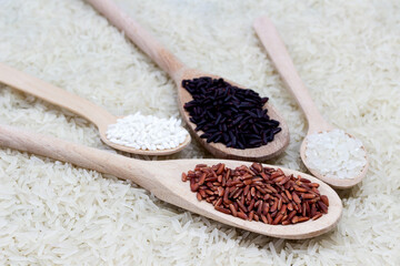 Red rice, glutinous rice, black basmati rice in wooden spoons. Basmati rice is scattered as food background