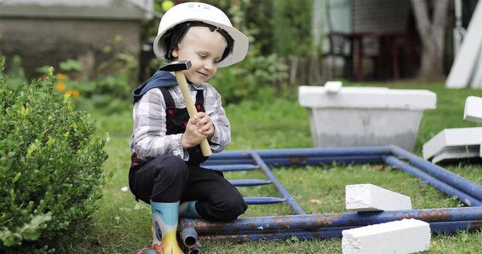Little builder in a white helmet knocks and hits a piece of styrofoam with a hammer. Happy little boy playing with tools on a construction site. Child helping parents to spike and nail.