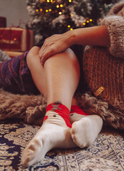 Woman sitting by the Christmas tree on a cozy boho rug wearing festive reindeer socks. Bokeh Christmas lights and presents in the background. Cozy holiday mood. Closeup of festive reindeer socks. 