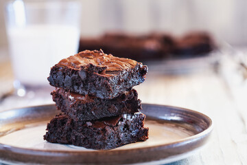 Fresh homemade fudgy brownies stacked on a saucer over a white rustic wooden table. Extreme shallow depth of field with blurred background and a glass of milk.