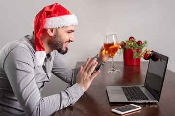 young man with Christmas hat toasting through laptop and cell phone celebrating Christmas online smiling and happy