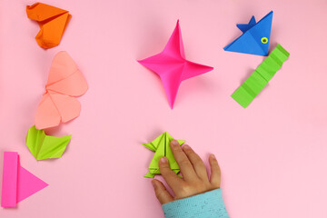 A child makes origami from colored paper on a pink background