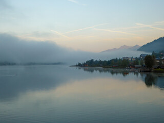 A foggy morning at the Weissensee lake in Austria. The lake is surrounded by high Alps. The calm surface of the lake reflects the forest. Sunbeams reaching the high peaks. Serenity and peacefulness