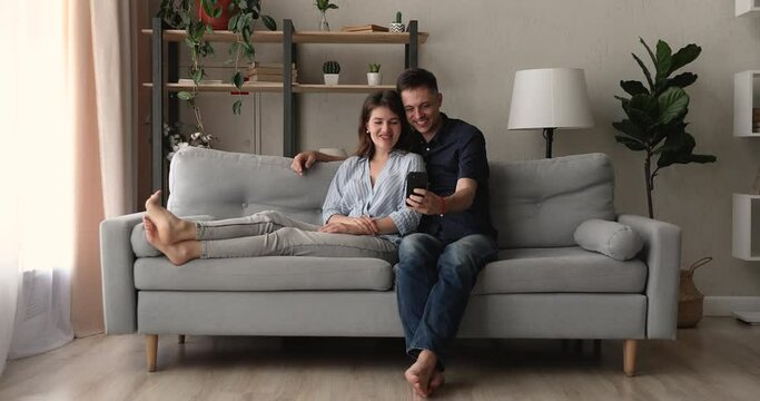 Millennial couple relaxing on couch taking selfie pictures using smartphone, having fun with new cool application, enjoy carefree weekend lazy day with wireless modern technology and internet concept