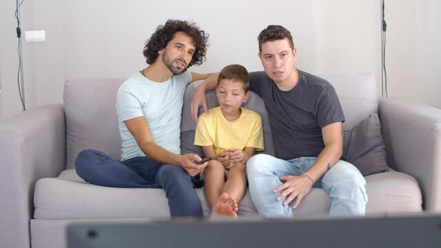 Two fathers and son watching movie on TV at home, sitting in living room together, using remote control and staring away. Family and home entertainment concept.