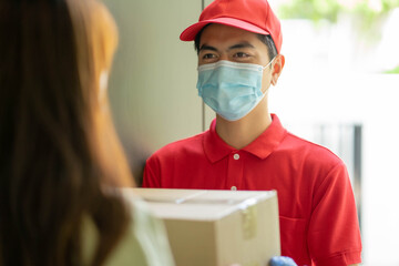 Deliver man wearing face mask in red uniform handing a parcel box over to a customer in front of...
