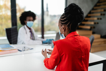 Female Afro American doctor talking with her patient behind protective glass. They wearing protective face masks as a virus pandemic protection. Coronavirus healthcare concept.