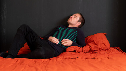 MAN IN HIS ROOM LYING ON THE BED WITH QUILT AND ORANGE PILLOWS AND BLACK WALL