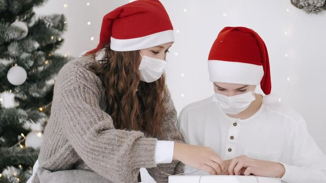 Christmas or New Year celebration. Covid-19 pandemic. The mother ans her son in party hats are unwrapping the gifts near Christas tree.