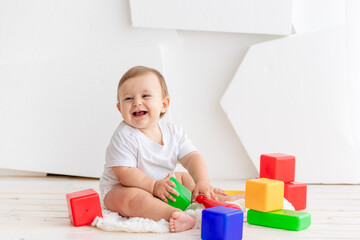 happy little baby six months old in a white t shirt and diapers playing at home on a Mat in a bright room with bright colored cubes