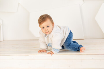 Naklejka premium a small child six months old in a knitted warm jacket and blue jeans crawls at home in a bright room