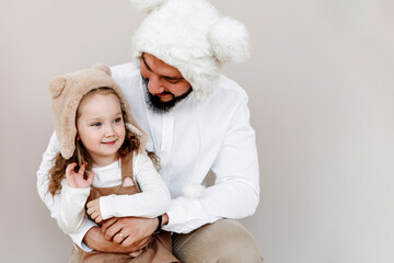 little girl in the arms of her dad. A man and his little daughter in large fur hats on a beige background. Family, holiday, christmas concept. Selective focus.