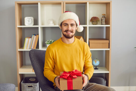 Young positive man in Christmas Satna hat sitting holding holiday present in hands