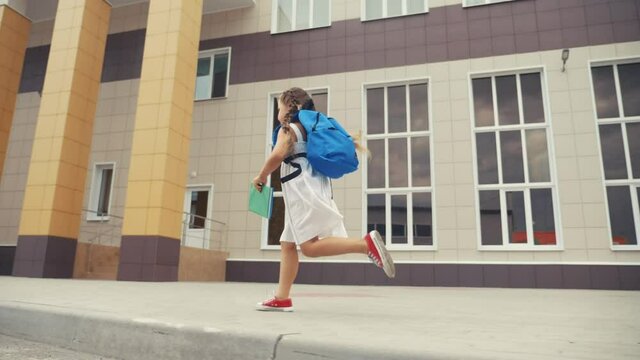 little girl kid with a backpack and a textbook runs hurrying to school. fun education concept. little schoolgirl with a backpack runs to the school building. child running with textbook back view