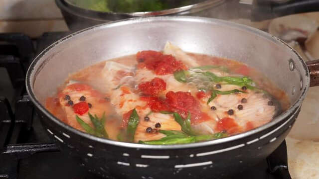 Woman's hand opens the lid of a frying pan with Salmon fish boiling in water with tarragon herbs, tomatoes and black pepper