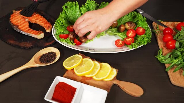 Woman's hand puts fried salmon on the plate with lettuce, potatoes and halfed cherry tomatoes