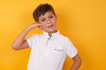 little cute Caucasian boy kid wearing white t-shirt against yellow wall imitates telephone conversation, makes phone call gesture with hands, has confident expression. Call me!