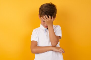 little cute Caucasian boy kid wearing white t-shirt against yellow wall making facepalm gesture while smiling amazed with stupid situation.