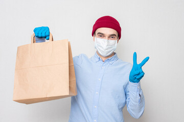Fototapeta na wymiar Courier in a medical mask and medical gloves holds a paper bag and symbol peace. Concept of safe delivery. Portrait, mockup