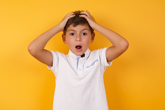 Horrible, stress, shock. Portrait emotional crazy little cute Caucasian boy kid wearing white t-shirt against yellow wall clasping head in hands. Emotions, facial expression concept.