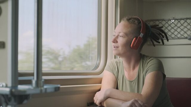Happy woman passenger rides on train and listens to music with headphones while sitting near window.